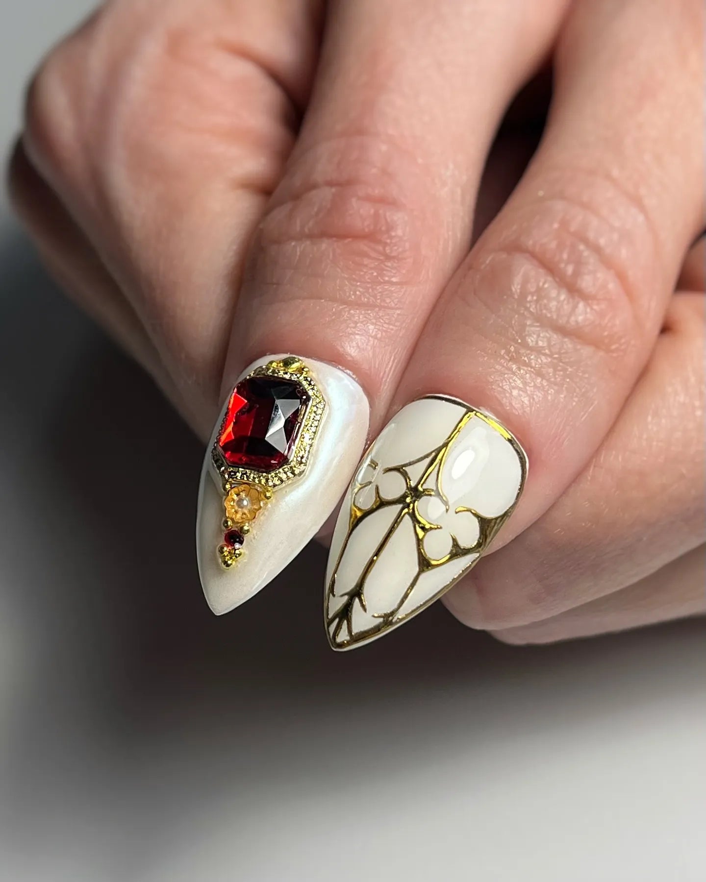 Hand holding handmade white stiletto press-on nails with gold accents, red gem and pearl decor