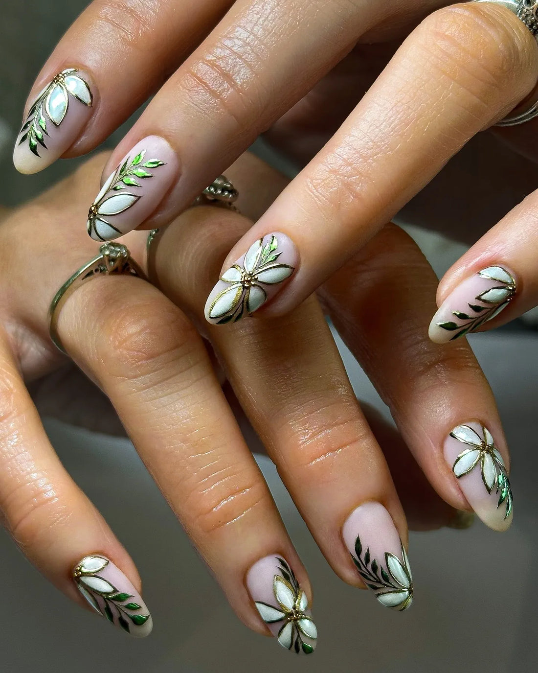 Hand with almond-shaped press-on nails featuring white pearl floral designs and metallic green leaves.