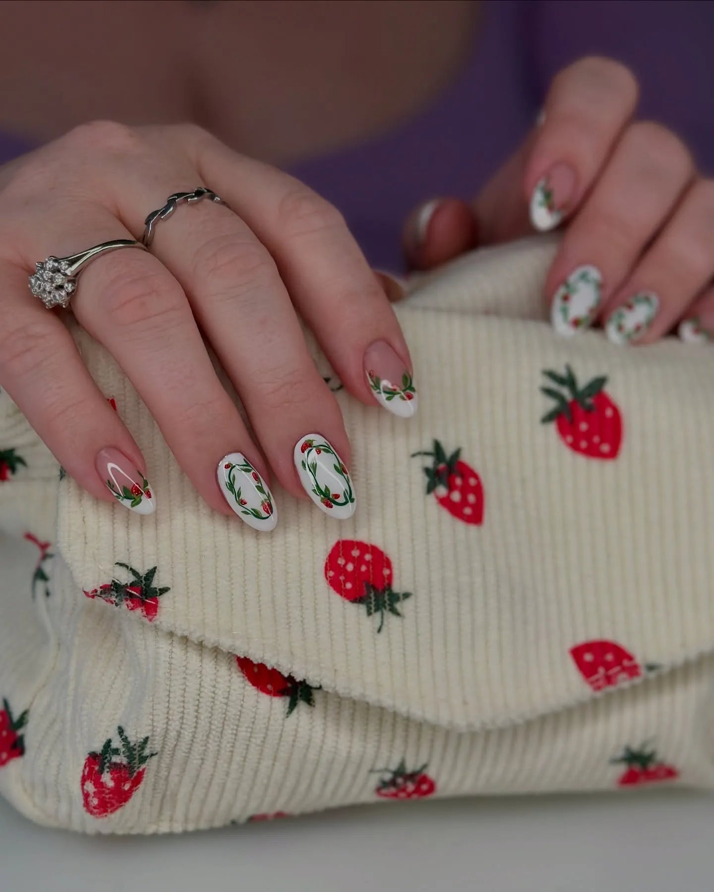 Hand with white artistic press-on nails featuring wreath and strawberry designs, holding a strawberry-patterned cream clutch.